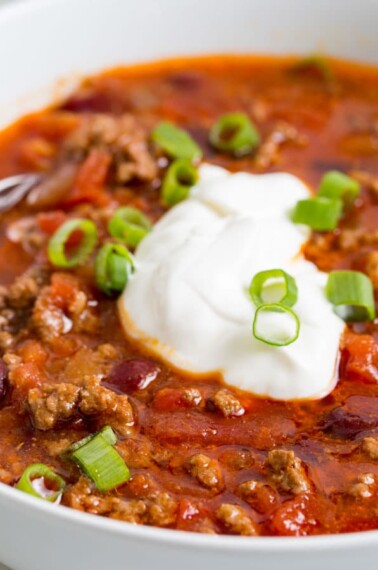 Image of easy slow cooker ground beef chili with beans in a white bowl, topped with sour cream and fresh scallions. A silver spoon is dipped in the bowl.
