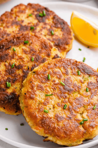 Three air fryer crab cakes on a plate with two lemon wedges.