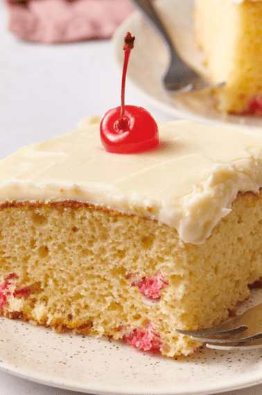 A slice of frosted cherry cake topped with a cherry on a plate.