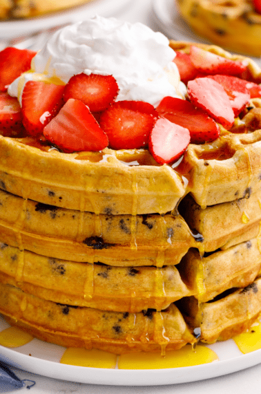A stack of four chocolate chip waffles topped with strawberries and whipped cream.