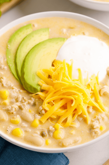 A bowl of white bean chicken chili topped with cheese, yogurt, and avocado slices.