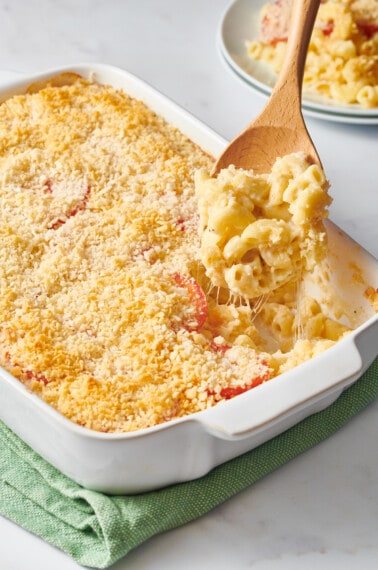 A wooden spoon lifting a scoop of Ina Garten mac and cheese from a baking dish.