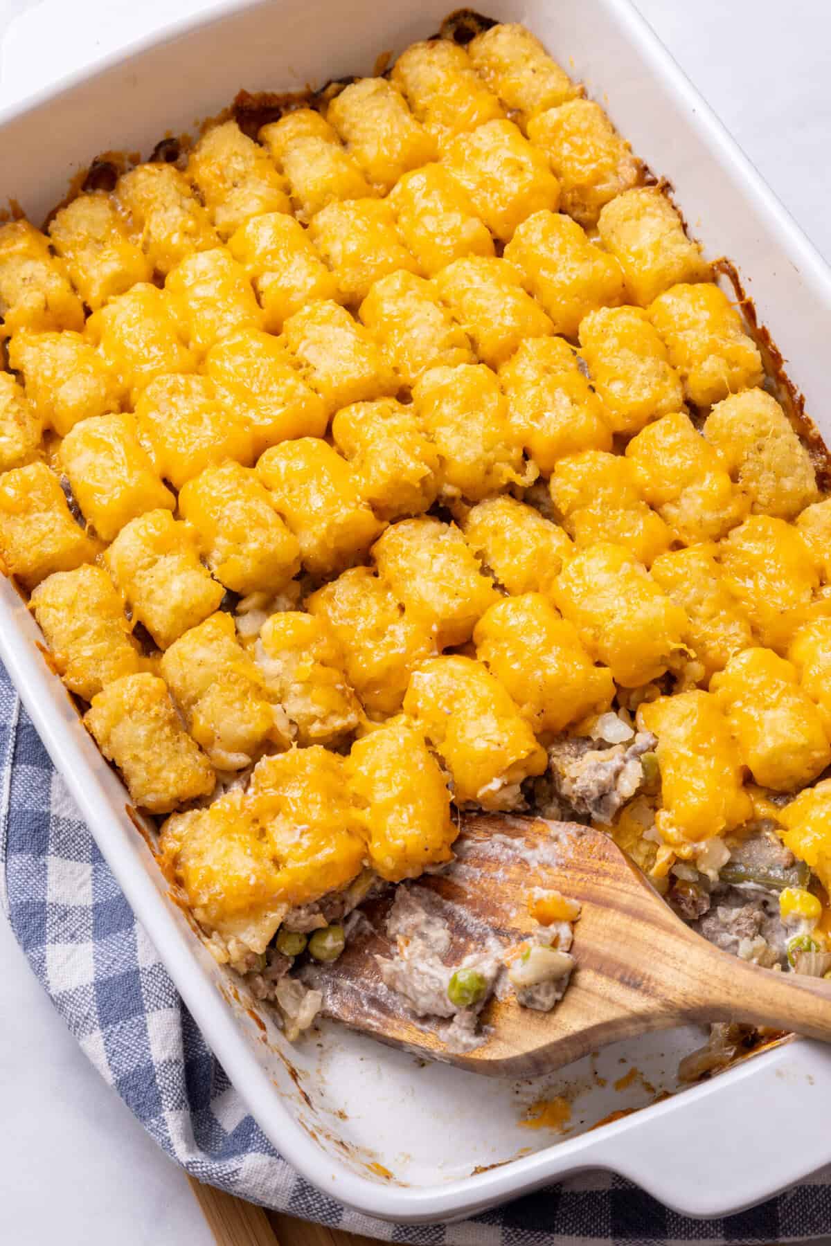 Tater Tot Casserole Tater Tot Casserole in a white baking dish being scooped out by a wooden spoon.